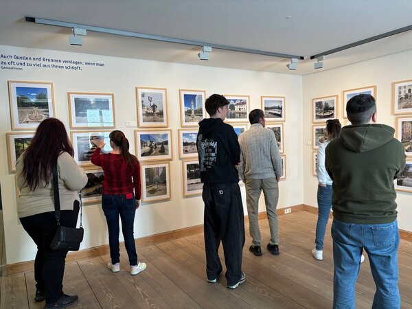 Eine Gruppe von Menschen betrachtet eine Fotoausstellung mit gerahmten Bildern von Brunnen und Wasserquellen. Ein Zitat an der Wand von Demosthenes warnt vor der Übernutzung von Wasserquellen: „Auch Quellen und Brunnen versiegen, wenn man zu oft und zu viel aus ihnen schöpft.“