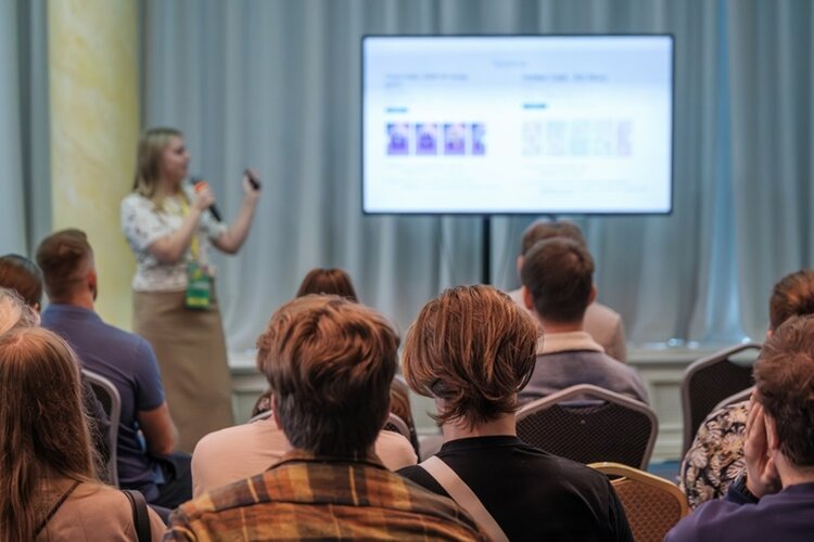 Mehrere Personen sitzen in einem Konferenzraum und blicken auf eine große Präsentationsleinwand. Eine Person steht vorn, hält einen Zeigestab oder Fernbedienung und spricht zu den Zuhörenden. Die Leinwand zeigt eine Präsentation mit Text und Bildern, die jedoch nicht lesbar sind. Die Szene wirkt wie eine Schulung oder ein Vortrag. Die Vorhänge im Hintergrund sind geschlossen, das Licht ist gedämpft.