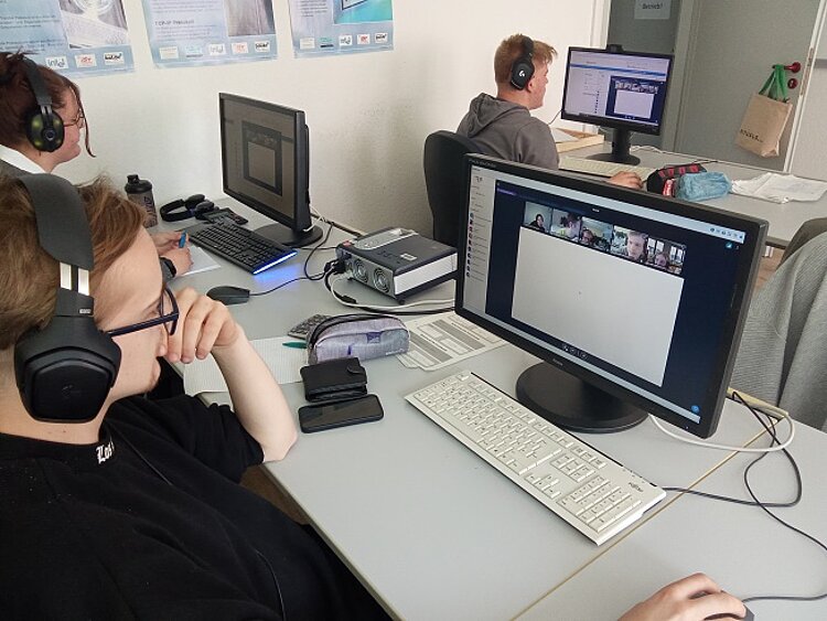 Bericht zur Berufsschulschließung am 08.05.2025 Detailaufnahme eines jungen Erwachsenen, der mit Kopfhörern vor einem Monitor sitzt und an einer Videokonferenz teilnimmt. Der Monitor zeigt die Teilnehmer einer Online-Konferenz. Auf dem Schreibtisch befinden sich persönliche Gegenstände wie Smartphone, Etui und technisches Zubehör. Im Hintergrund arbeiten weitere Personen ebenfalls an Computern.