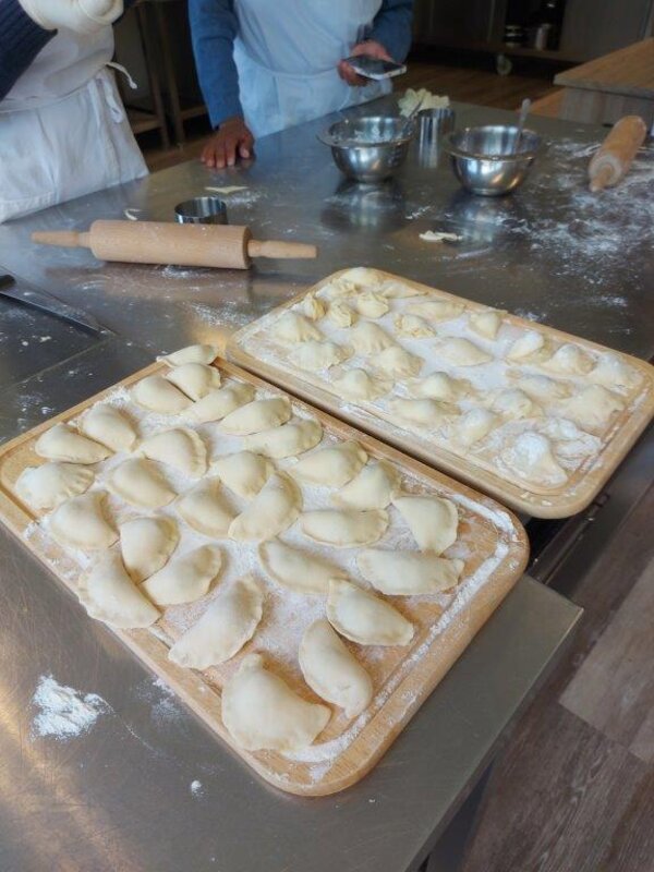Frisch zubereitete Teigtaschen liegen bemehlt auf zwei Holzbrettchen, bereit zum Kochen. Im Hintergrund liegen Nudelholz, Metallschüsseln und weitere Backutensilien auf einem Edelstahltisch.