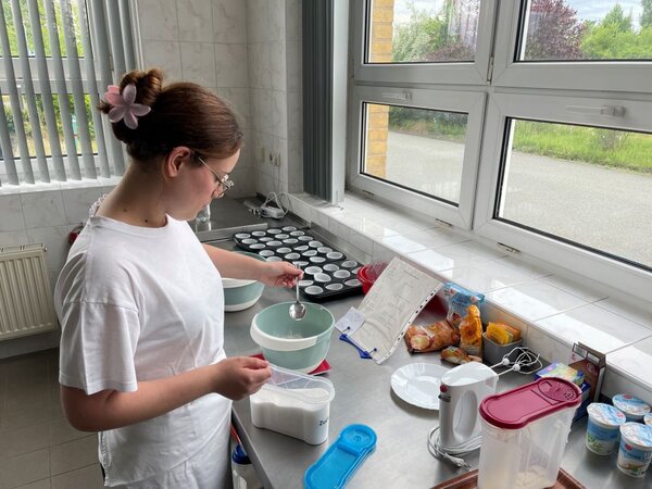  Eine Person in weißer Kleidung und mit einer pinken Haarspange beugt sich über einen Küchentisch. Sie hält einen Löffel in der Hand und füllt Zucker in eine Schüssel. Im Hintergrund sind Backbleche mit Muffinförmchen, eine Küchenmaschine und weitere Zutaten zu sehen.