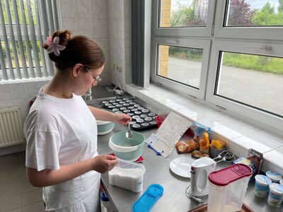  Eine Person in weißer Kleidung und mit einer pinken Haarspange beugt sich über einen Küchentisch. Sie hält einen Löffel in der Hand und füllt Zucker in eine Schüssel. Im Hintergrund sind Backbleche mit Muffinförmchen, eine Küchenmaschine und weitere Zutaten zu sehen.