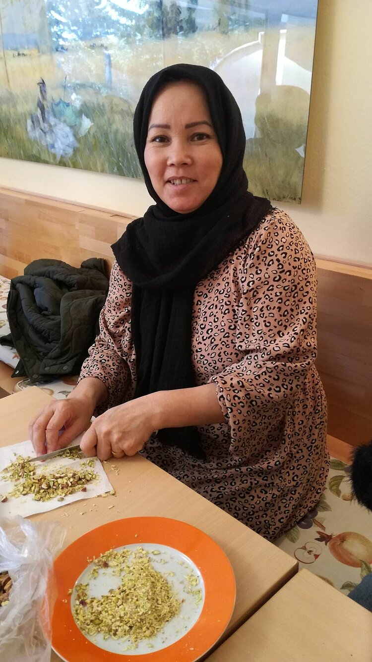 Eine Frau mit schwarzem Kopftuch und einem beigen Kleid mit Leopardenmuster sitzt an einem Tisch und hackt Pistazien auf einem Schneidebrett. Vor ihr steht ein Teller mit bereits zerkleinerten Nüssen.