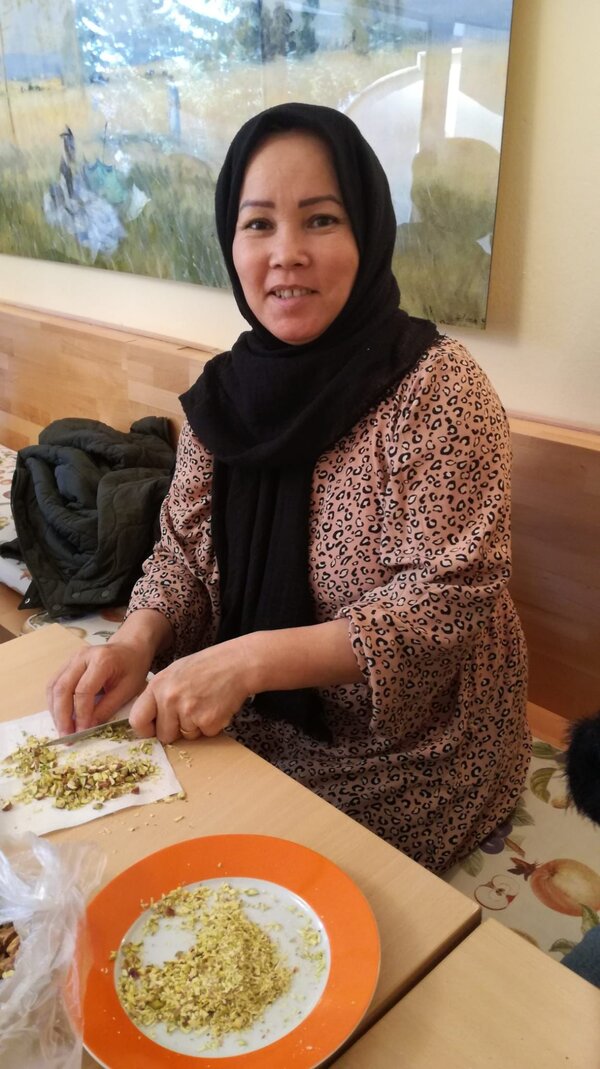 Eine Frau mit schwarzem Kopftuch und einem beigen Kleid mit Leopardenmuster sitzt an einem Tisch und hackt Pistazien auf einem Schneidebrett. Vor ihr steht ein Teller mit bereits zerkleinerten Nüssen.