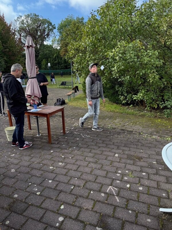 Im Vordergrund steht ein junger Mann mit Basecap, grauer Jacke und heller Hose auf einem gepflasterten Platz. Er blickt nach oben in Richtung eines weißen Papierfliegers, der in der Luft schwebt. Links daneben steht ein Holztisch mit einem geschlossenen Sonnenschirm und mehreren Gegenständen darauf. Im Hintergrund sind weitere Personen, ein grüner Zaun und Bäume zu sehen. Die Szene spielt im Freien bei Tageslicht.