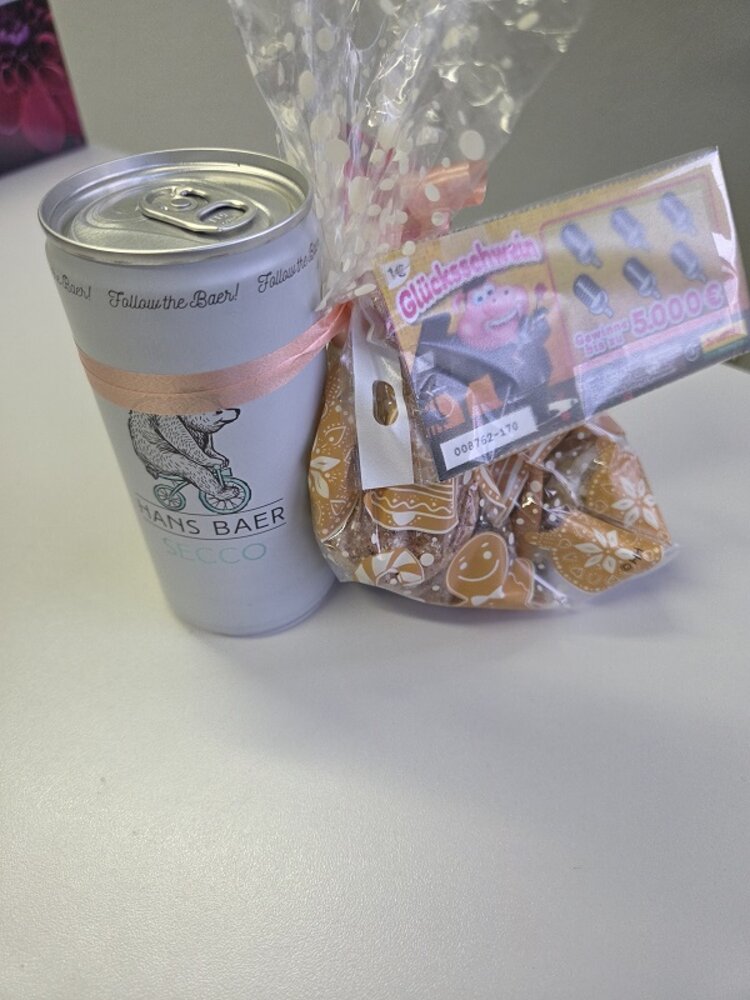 Ein kleines Geschenkset auf einem Tisch: ein Becher mit Bärenlogo, eine transparente Tüte mit verpackten Keksen und ein Lottolos, dekorativ mit Schleife zusammengebunden.