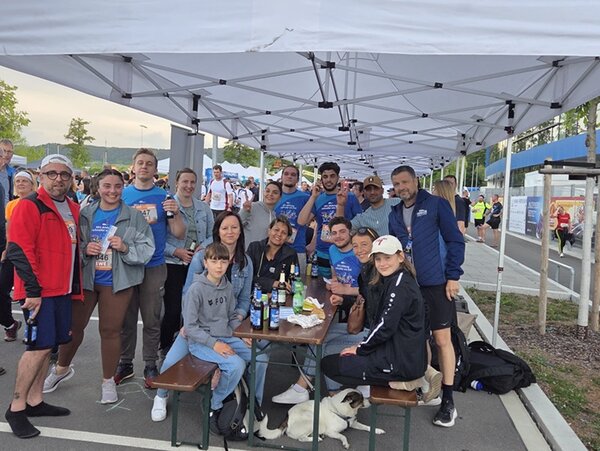 Die Teilnehmenden der Euro Akademie und der Euro-Schulen Jena sitzen gemeinsam auf einer Bank, darunter liegt ein Hund, einige Teilnehmende stehen um die Bank und den Tisch herum, und posieren für ein Foto. Sie alle sind sportlich für den 14. Jenaer Firmenlauf gekleidet. 