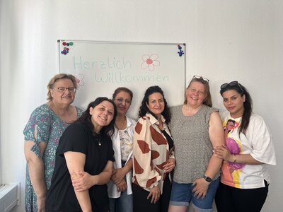Sechs Frauen stehen nebeneinander vor einem weißen Whiteboard an den Euro-Schulen Hannover. Sie lächeln und blicken in die Kamera. Das Whiteboard ist mit bunten Filzstiften beschriftet und zeigt die Worte 'Herzlich Willkommen' sowie zwei gezeichnete Blumen. Die Frauen tragen unterschiedliche, überwiegend sommerliche Kleidung. Die Szene ist freundlich und einladend.