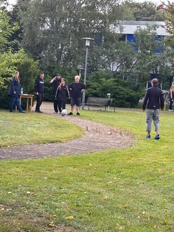 Mehrere Personen stehen auf einer Wiese und einem geschwungenen Weg in einem Park. Im Vordergrund sind auf dem Weg Holzklötze in Reihen aufgestellt. Eine Person steht mit dem Rücken zur Kamera und blickt auf die Gruppe. Im Hintergrund sind Bäume, Sträucher, eine Bank und ein modernes Gebäude mit großen Fenstern zu sehen. Die Szene wirkt entspannt und ungezwungen.