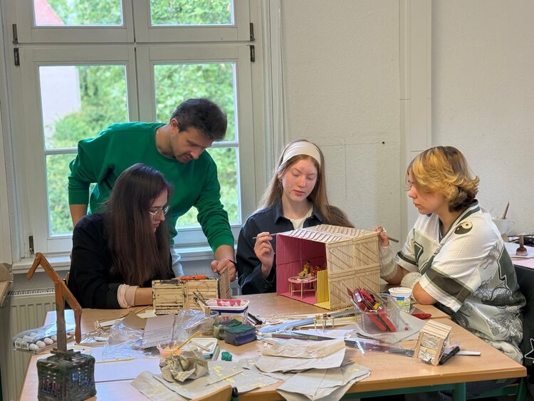 Vier Personen arbeiten gemeinsam an kleinen Modellhäusern an einem Tisch in einem hellen Klassenraum. Vor ihnen liegen Bastelmaterialien.