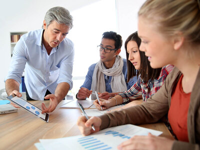 Eine Gruppe von vier Personen sitzt an einem Tisch und diskutiert über Dokumente und Diagramme. Ein Mann mit grauen Haaren zeigt auf ein Tablet, während die anderen drei Personen, zwei Frauen und ein Mann, Notizen machen und aufmerksam zuhören.
