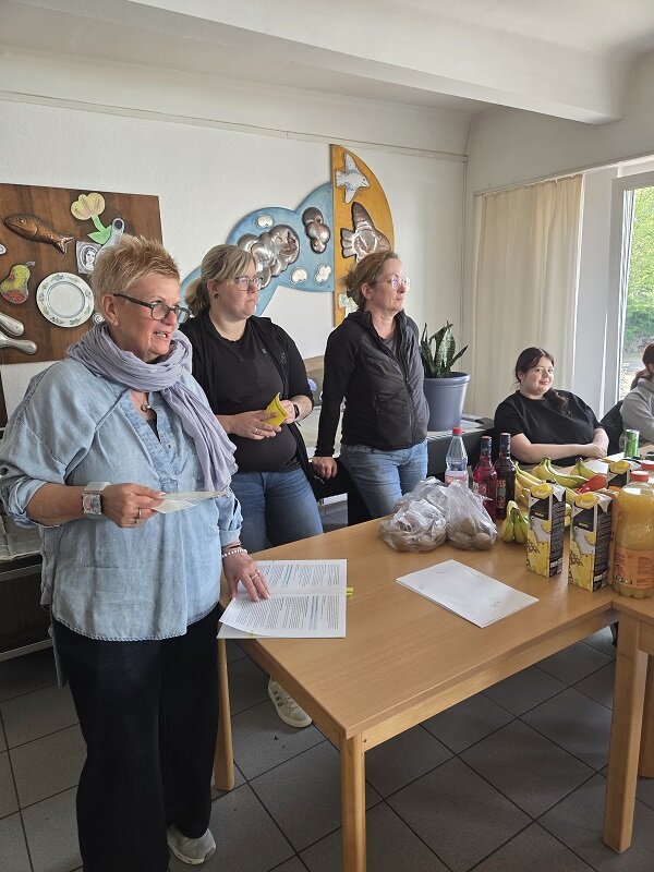 Drei Personen, möglicherweise die Projektleiterinnen, stehen in einem Raum mit hellen Wänden und großen Fenstern. Auf dem Tisch vor ihnen liegen verschiedene Lebensmittel wie Bananen, Kiwis in Tüten und Kartons mit Saft. Eine Person hält ein Blatt Papier in der Hand. Im Hintergrund sitzen zwei Personen am Tisch.