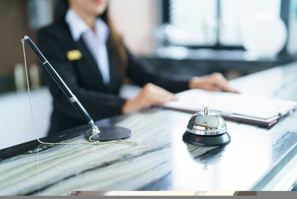 Zu sehen ist eine Rezeption eines Hotels. Auf dem Dresen steht eine Klingel und es steht dort ein Kugelschreiber. Im Hintergrund steht eine Frau in schwarzem Anzug.