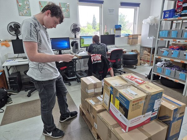 Zwei Auszubildende im Lagerbüro: Einer prüft eine Lieferliste vor einem Stapel Pakete, der andere arbeitet am Computer.