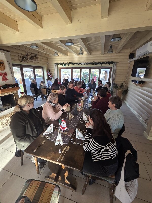 Viele Menschen sitzen an einer langen Tafel in einem hellen, holzverkleideten Raum und unterhalten sich bei einem gemeinsamen Essen.