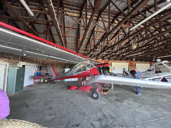 Blick in einen Hangar mit einem rot-silbernen Motorflugzeug und einem weiteren Flugzeug im Hintergrund; mehrere Personen halten sich zwischen den Maschinen auf.