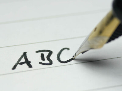 Ein Füllfederhalter schreibt die Buchstaben 'ABC' auf ein Blatt Papier. Der Füllfederhalter hat eine schwarze Feder und einen goldfarbenen Griff. Das Papier ist weiß und hat feine Linien, die als Schreibhilfe dienen.