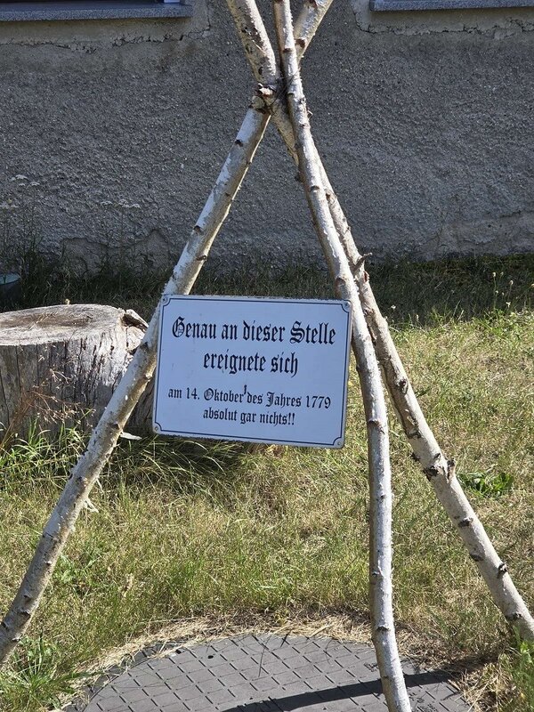Ein weißes Schild mit schwarzer Schrift hängt an einem Holzgestell aus Ästen. Der Text auf dem Schild lautet: 'Genau an dieser Stelle ereignete sich am 14. Oktober des Jahres 1779 absolut gar nichts!!'. Im Hintergrund sind ein Baumstumpf und eine graue Wand zu sehen.