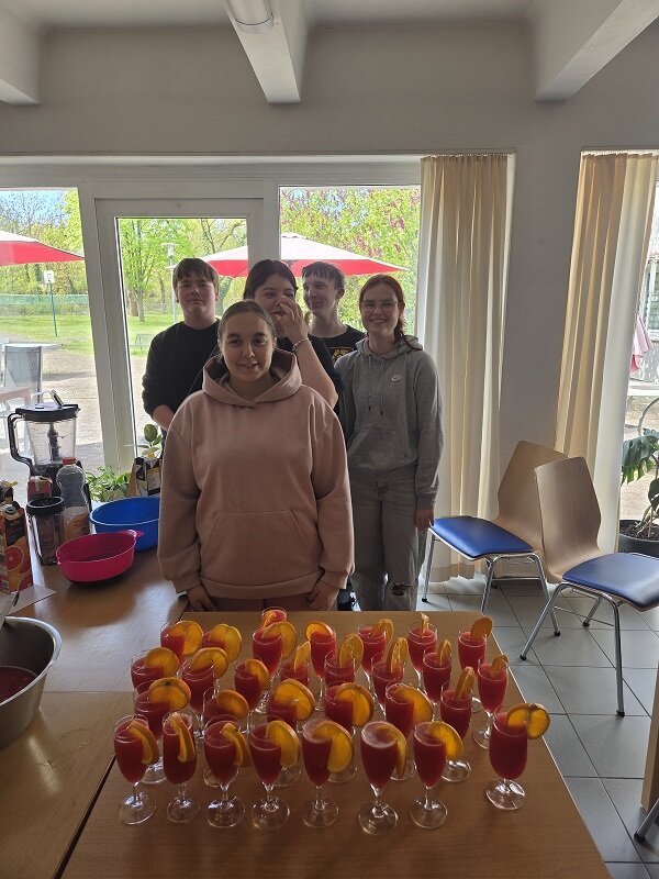 Fünf Personen stehen hinter einem Tisch, auf dem zahlreiche Gläser mit einem roten Getränk und Orangenscheiben als Dekoration arrangiert sind. Die Gruppe posiert lächelnd für das Foto in einem hellen Raum mit großen Fenstern.