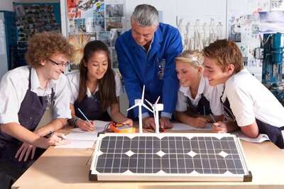 Ein Ausbilder erklärt seinen Auszubildenden etwas über Windenergie und Solarpaneele in einer Berufsschule. Die weiblichen und männlichen Azubis beugen sich interessiert und gut gelaunt über das aufgebaute Model.