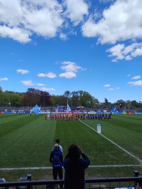 Zwei Fußballmannschaften stehen in einer Reihe auf einem Rasenplatz, jeweils in farblich unterschiedlichen Trikots. Im Hintergrund sind große Fahnen zu sehen, die von Personen gehalten werden. Der Himmel ist blau mit vereinzelten weißen Wolken. Im Vordergrund stehen zwei Personen am Spielfeldrand, eine davon hält ein Mobiltelefon oder eine Kamera. Im Hintergrund sind Bäume und eine Tribüne mit Zuschauern erkennbar.