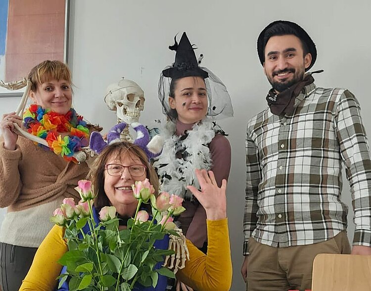 Vier Personen in festlichen Verkleidungen posieren in einem Raum. Eine Frau mit Blumenkranz und einem Skelettkopf in der Hand, eine andere mit Hexenhut und Federboa, ein Mann mit einem Halstuch sowie eine ältere Dame mit violetten Plüsch-Ohren und einem Strauß rosa Rosen. Alle lachen fröhlich in die Kamera.