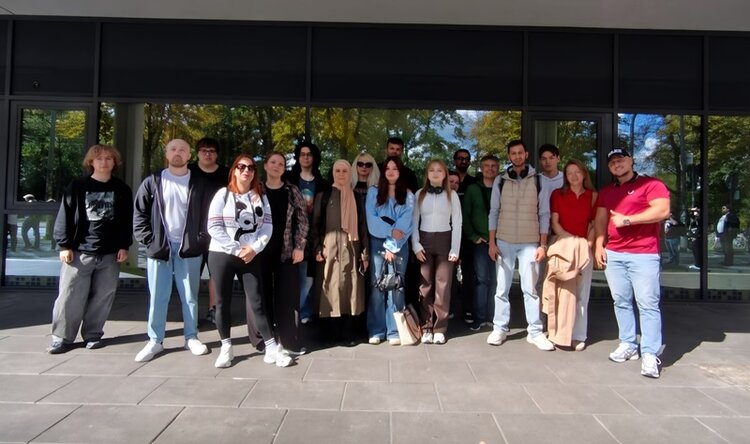 Die Gruppe der Teilnehmenden steht nebeneinander auf einem gepflasterten Bereich vor einer Glasfassade. Die Personen tragen unterschiedliche Freizeitkleidung, darunter Jacken, Hosen und T-Shirts. Im Hintergrund spiegeln sich Bäume und Himmel in den Fenstern. Die Szene ist von Tageslicht erhellt.