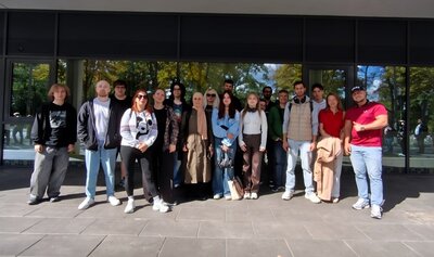 Die Gruppe der Teilnehmenden steht nebeneinander auf einem gepflasterten Bereich vor einer Glasfassade. Die Personen tragen unterschiedliche Freizeitkleidung, darunter Jacken, Hosen und T-Shirts. Im Hintergrund spiegeln sich Bäume und Himmel in den Fenstern. Die Szene ist von Tageslicht erhellt.