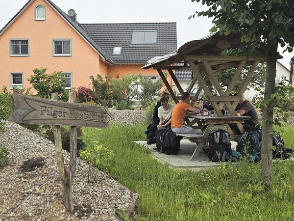 Wandernde rasten mit ihren Rucksäcken an einem überdachten Picknicktisch neben einem Holzschild mit der Aufschrift „Pilger Raststätte“.