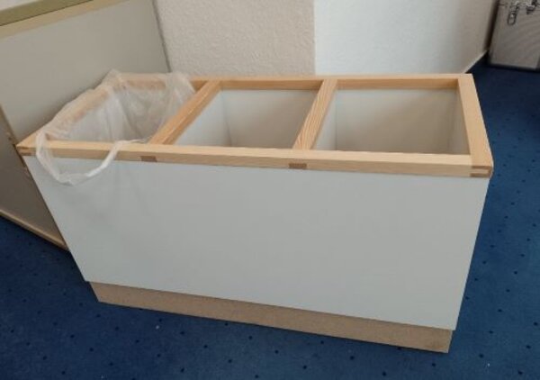 Ein rechteckiger Mülltrennungsbehälter aus Holz und weißen Seitenwänden, bestehend aus drei Fächern. Links ist ein Fach mit einem Müllbeutel ausgestattet, während die anderen beiden Fächer leer sind. Der Behälter steht auf einem blauen Teppichboden und ist vor einer weißen Wand platziert.