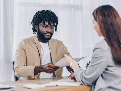 Ein Mann mit Dreadlocks und einem hellbraunen Blazer sitzt an einem Schreibtisch und reicht einer Frau in einem grauen Anzug ein Dokument. Beide wirken professionell und sind in ein Gespräch vertieft. Auf dem Tisch liegen verschiedene Unterlagen und ein Laptop.