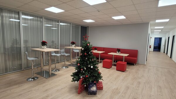 Moderner Aufenthaltsbereich mit Stehtischen und Barhockern, roter Sitzbank und in der Mitte ein geschmückter Weihnachtsbaum mit Geschenken darunter.