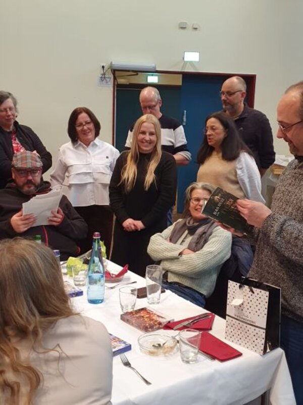 Mehrere Menschen stehen und sitzen dicht beieinander um einen gedeckten Tisch und schauen gemeinsam in Karten oder Hefte. Einige lächeln, andere lesen konzentriert, auf dem Tisch stehen Gläser, eine Wasserflasche und kleine Geschenke.