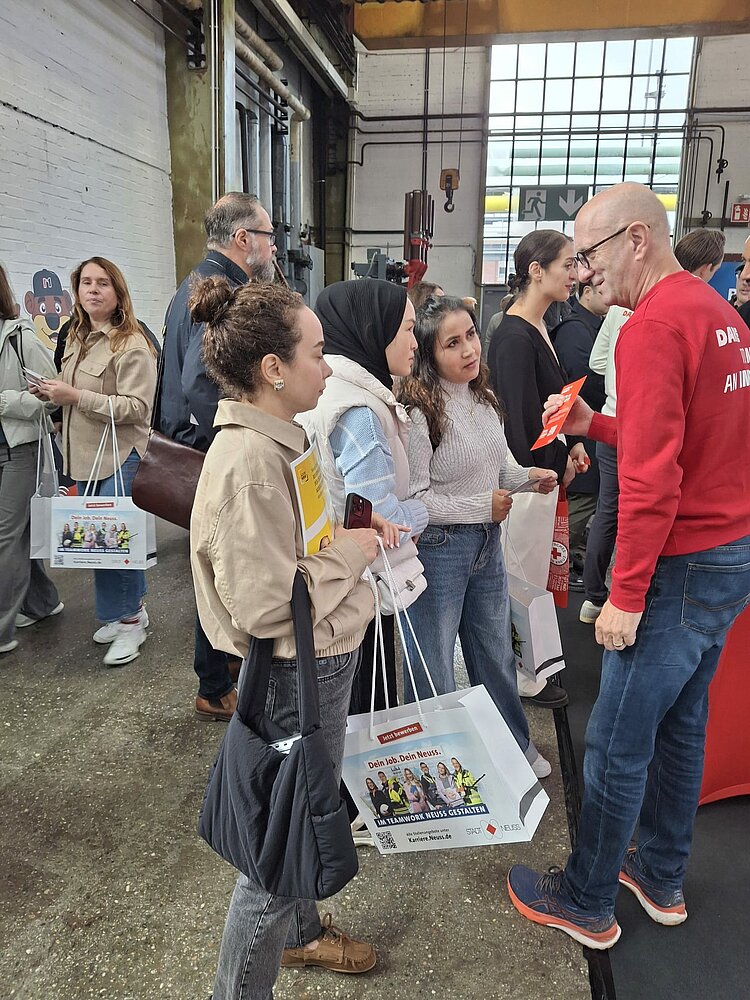 Ein Mitarbeiter an einem Messestand hält den Maßnahmenteilnehmerinnen einen Flyer hin und informiert über die freien Stellen.