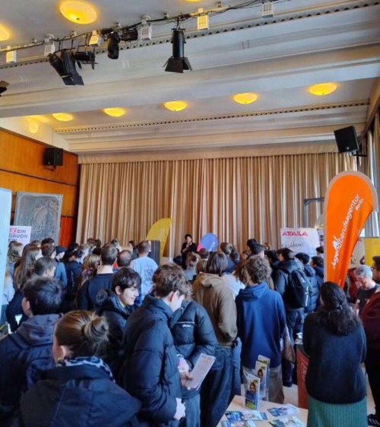 Ein großer Raum mit vielen jungen Menschen, die sich an verschiedenen Infoständen über Bildungs- und Berufsmöglichkeiten informieren. Auf einer kleinen Bühne spricht eine Person vor dem Publikum, während bunte Banner und Informationsmaterialien die Veranstaltung begleiten.