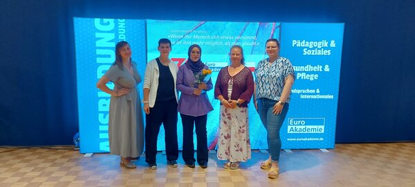 Fünf Frauen stehen nebeneinander auf einem Parkettboden vor einem großen, beleuchteten Hintergrund mit der Aufschrift "Euro Akademie" und den Bereichen "Pädagogik & Soziales", "Gesundheit & Pflege" sowie "Fremdsprachen & Internationales". Die Frau in der Mitte hält einen Blumenstrauß. Alle Personen blicken in die Kamera und lächeln.