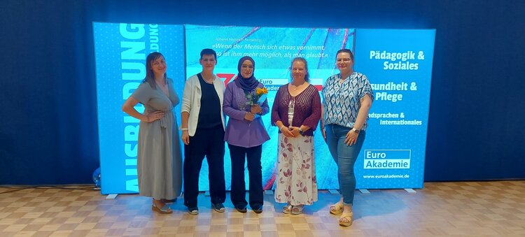 Fünf Frauen stehen nebeneinander auf einem Parkettboden vor einem großen, beleuchteten Hintergrund mit der Aufschrift "Euro Akademie" und den Bereichen "Pädagogik & Soziales", "Gesundheit & Pflege" sowie "Fremdsprachen & Internationales". Die Frau in der Mitte hält einen Blumenstrauß. Alle Personen blicken in die Kamera und lächeln.