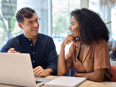 Zwei Personen sitzen an einem Tisch in einem hellen Raum, der wie ein Büro oder ein Café aussieht. Die Person links, ein Mann mit einer Brille und einem dunklen Hemd, arbeitet an einem Laptop. Die Person rechts, eine Frau mit lockigem Haar und einem braunen Blazer, scheint zuzuhören oder sich zu unterhalten. Beide lächeln und wirken entspannt und engagiert in ihrem Gespräch oder ihrer Arbeit.