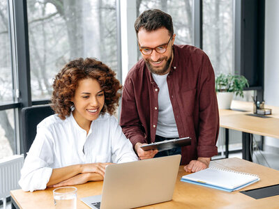 Zwei Kollegen arbeiten gemeinsam im Büro: Eine Person sitzt lächelnd am Laptop, während eine andere Person ihr freundlich über die Schulter schaut und ein Tablet in der Hand hält. Im Hintergrund sind große Fenster mit Blick auf Bäume zu sehen, und auf dem Tisch liegen ein Notizbuch und ein Glas Wasser.