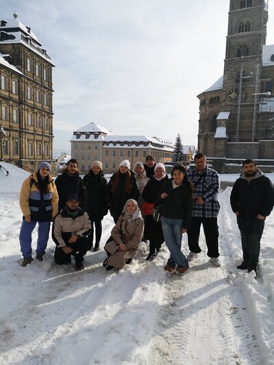 Eine Gruppe von Teilnehmenden der Euro-Schulen Bamberg posiert in einer verschneiten Landschaft für ein Foto. Sie stehen vor historischen Gebäuden, eines davon mit einem Glockenturm. Alle sind warm in Winterkleidung gekleidet, mit Mänteln, Mützen und Handschuhen. Der Boden ist schneebedeckt, der Himmel klar.