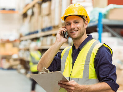 Eine Person trägt einen gelben Schutzhelm und eine hochsichtbare Sicherheitsweste, hält ein Klemmbrett und spricht am Mobiltelefon. Der Hintergrund scheint eine industrielle oder Lagerumgebung zu sein, mit Regalen, die mit verschiedenen Gegenständen gefüllt sind. Das Gesicht der Person ist aus Datenschutzgründen unscharf.
