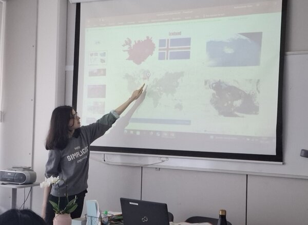 Eine junge weibliche Teilnehmerin mit braunen, schulterlangen Haaren steht vor am Whiteboard im Unterrichtsraum und hält ihre Präsentation vor ihren Mitteilnehmenden des B2-Sprachkurses an den Euro-Schulen Magdeburg und zeigt dabei mit dem Finger auf eine Karte.