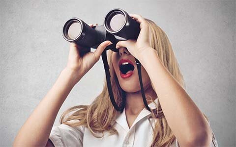 Eine Person mit langen blondem Haar und roten Lippenstift hält ein Fernglas vor ihre Augen. Sie hat den Mund staunend geöffnet.