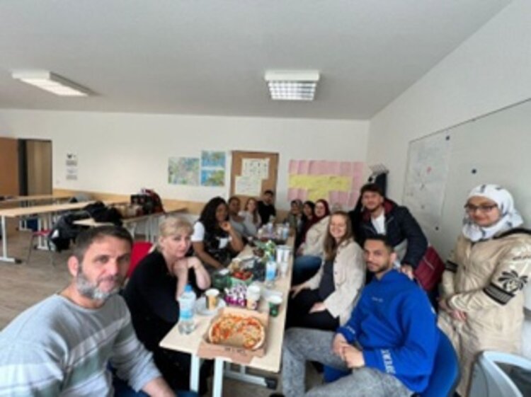 Mitarbeitende und Teilnehmende der Euro-Schulen Potsdam sitzen gemeinsam an einem gedeckten Tisch und verabschieden ihre Kollegin in den Mutterschutz.