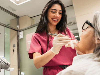 Eine lächelnde Zahnärztin in rosa Kittel behandelt eine Patientin mit Brille.