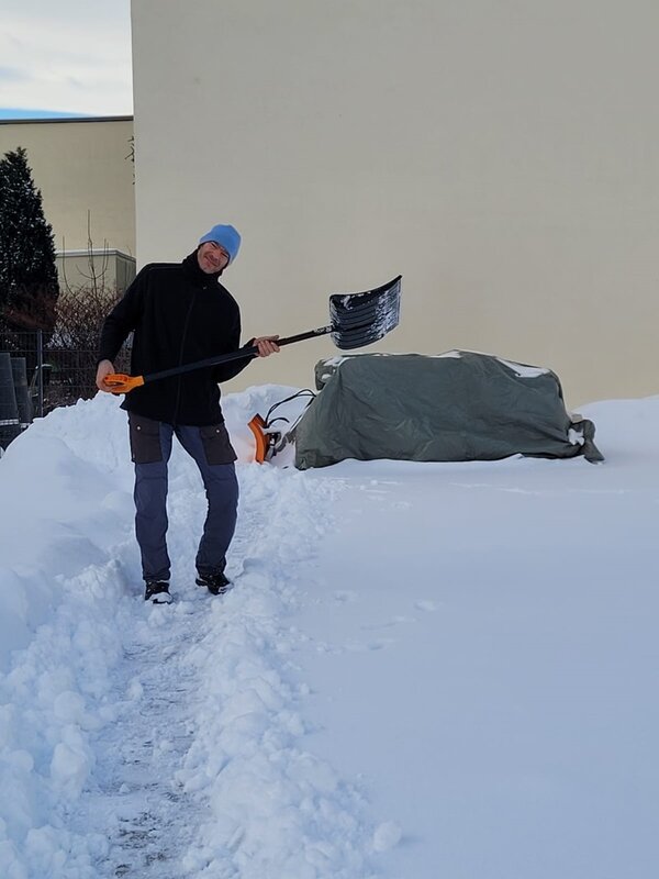 Der Hausmeister steht auf einer schneebedeckten Fläche und hält eine Schneeschaufel mit orangefarbenem Griff. Im Hintergrund ist eine große, helle Wand zu sehen. Ein mit einer grauen Plane abgedecktes Objekt sowie ein orangefarbenes Gerät liegen am Boden. Die Szene zeigt eine Schneeräumung, die laut Kontext vom Bauhof Hohenstein-Ernstthal zur Unterstützung der Euro-Schulen Hohenstein-Ernstthal durchgeführt wird.
