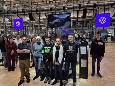 Teilnehmende und Lehrkraft posieren in der Gläsernen Manufaktur vor VW-Autos für ein Gruppenbild.