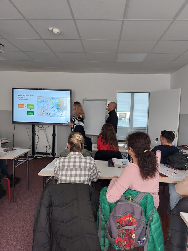 Eine Frau und ein Mann stehen vor einem großen Bildschirm und präsentieren Informationen zum Thema Ausbildungsvergütung. Die Zuhörer sitzen an ihren Plätzen und schauen auf den Bildschirm.