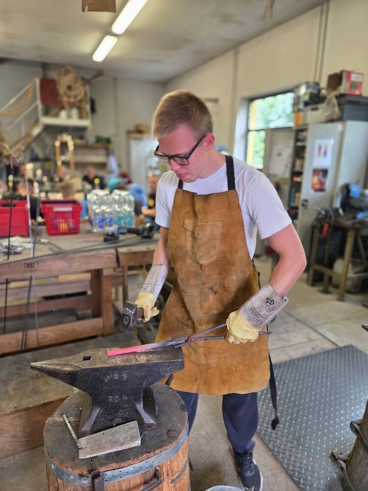 Jugendlicher mit Schutzbrille, Handschuhen und Schürze hämmert in einer Werkstatt auf erhitztes Metall auf einem Amboss.