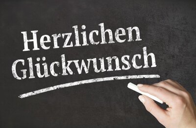 Eine Hand schreibt mit weißer Kreide die Worte 'Herzlichen Glückwunsch' auf eine schwarze Tafel.